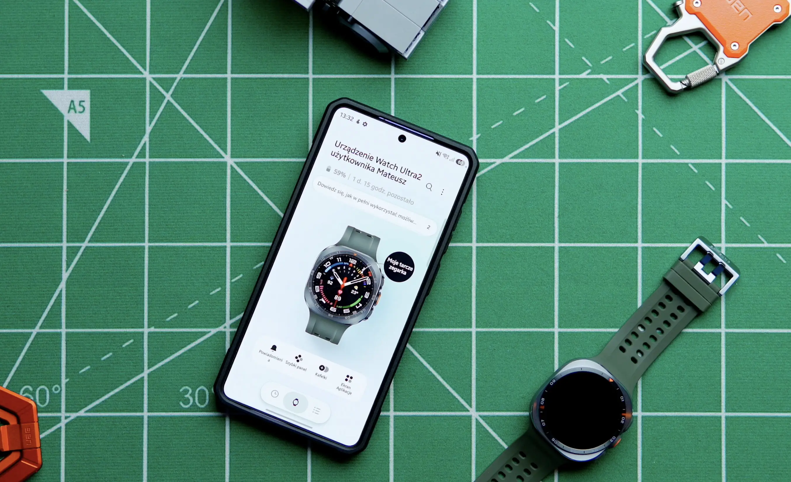 Smartwatch Samsung Galaxy Watch Ultra 2 leżący obok telefonu Samsunga pokazującego aplikację Wear..